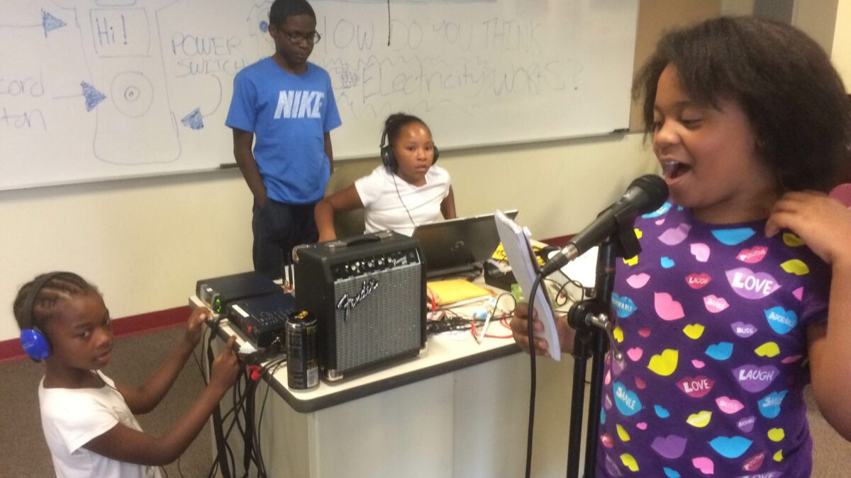 IMG_3327 A youth holding a notebook and speaking into a microphone as another youth wearing headphones listens and two youth, one wearing headphones, sit behind a laptop computer in a classroom