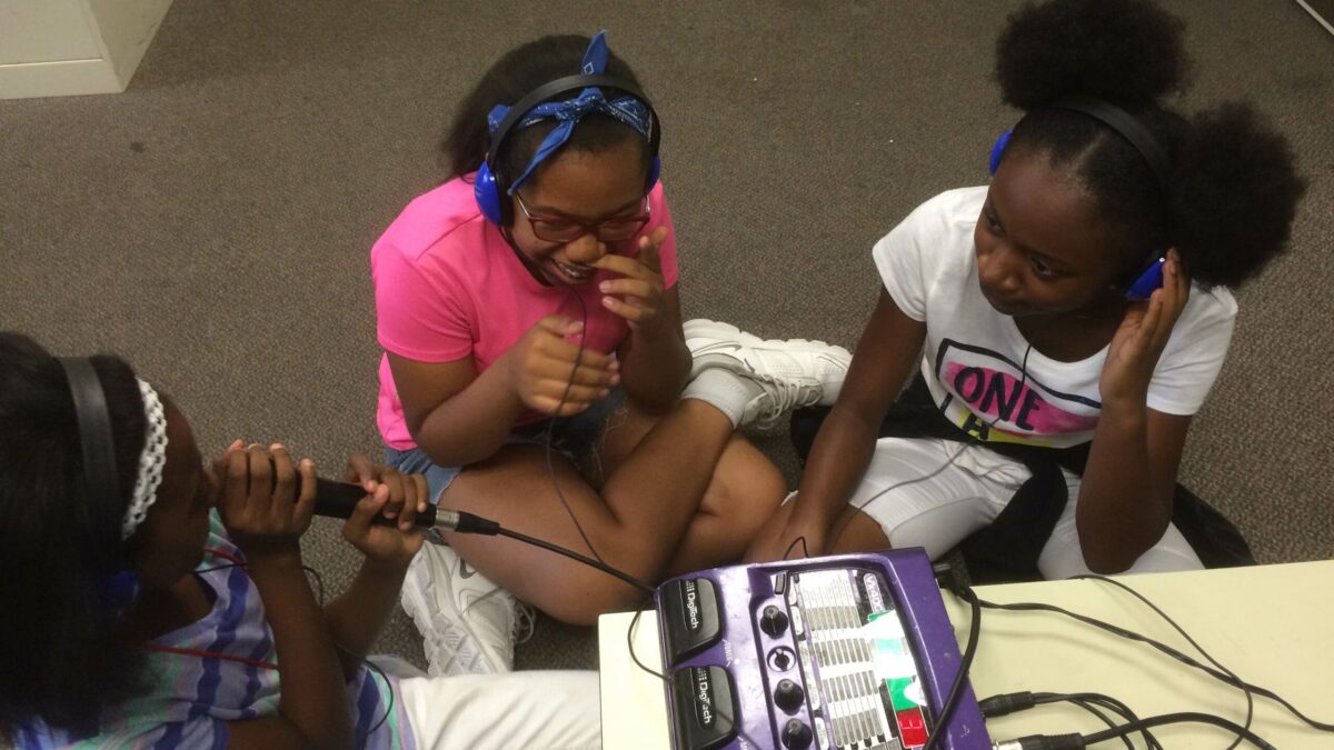 IMG_3431 Three youth wearing headphones smiling sitting around a mixer, one holding a microphone