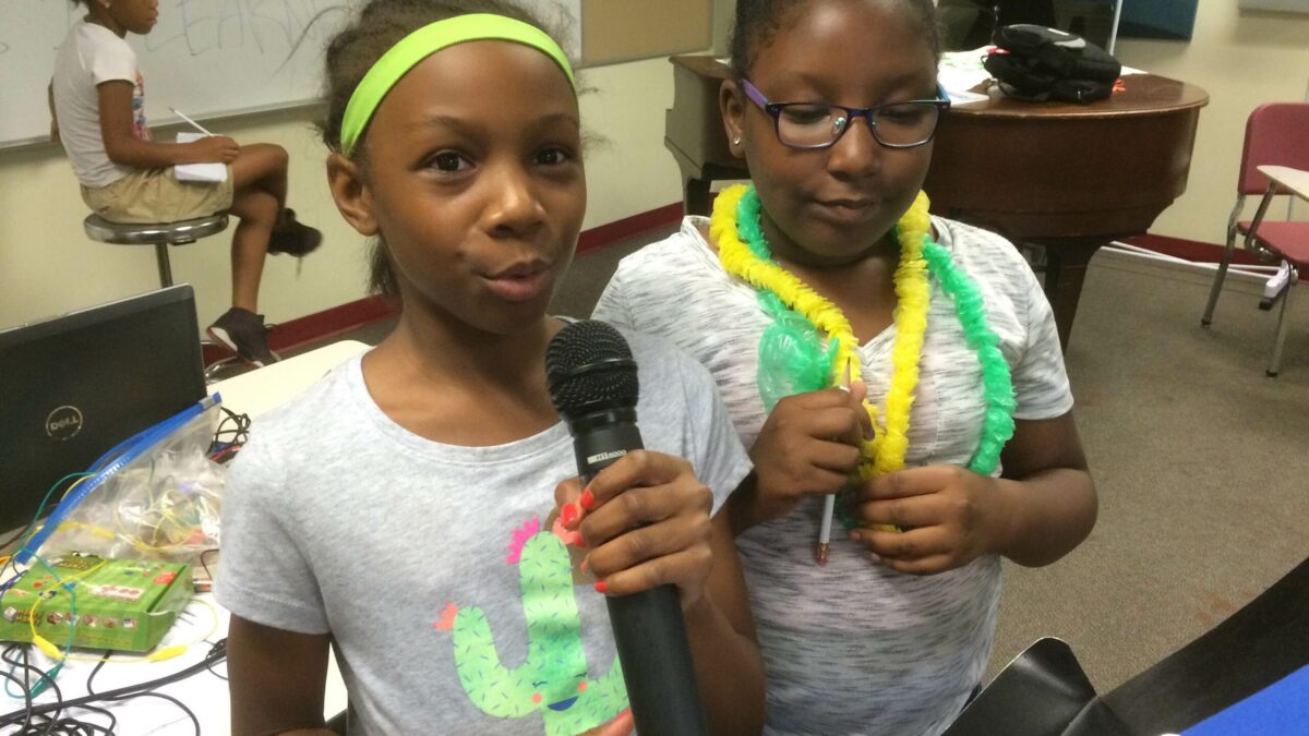 A youth holding and speaking into a microphone, a youth standing and looking down at a music stand, and a youth sitting in the background next to a whiteboard