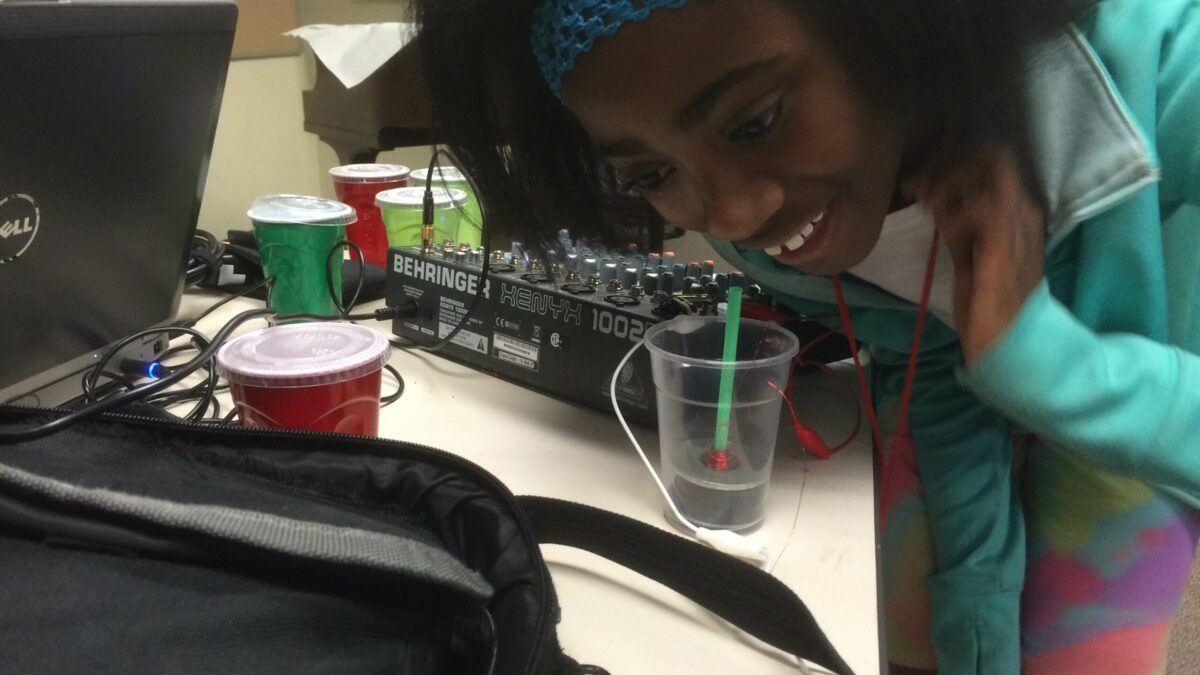 A youth crouched over a mixer speaking into a plastic cup with a thin cylinder inside