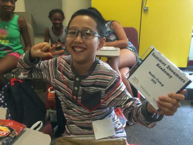 A youth flashing a piece sign and smiling at the camera while holding a white cd case