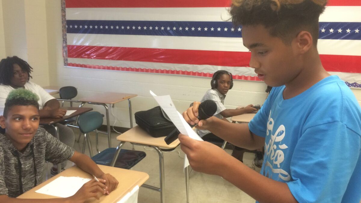 A youth reading a piece of paper holding a microphone as three youth listen sitting at classroom desks