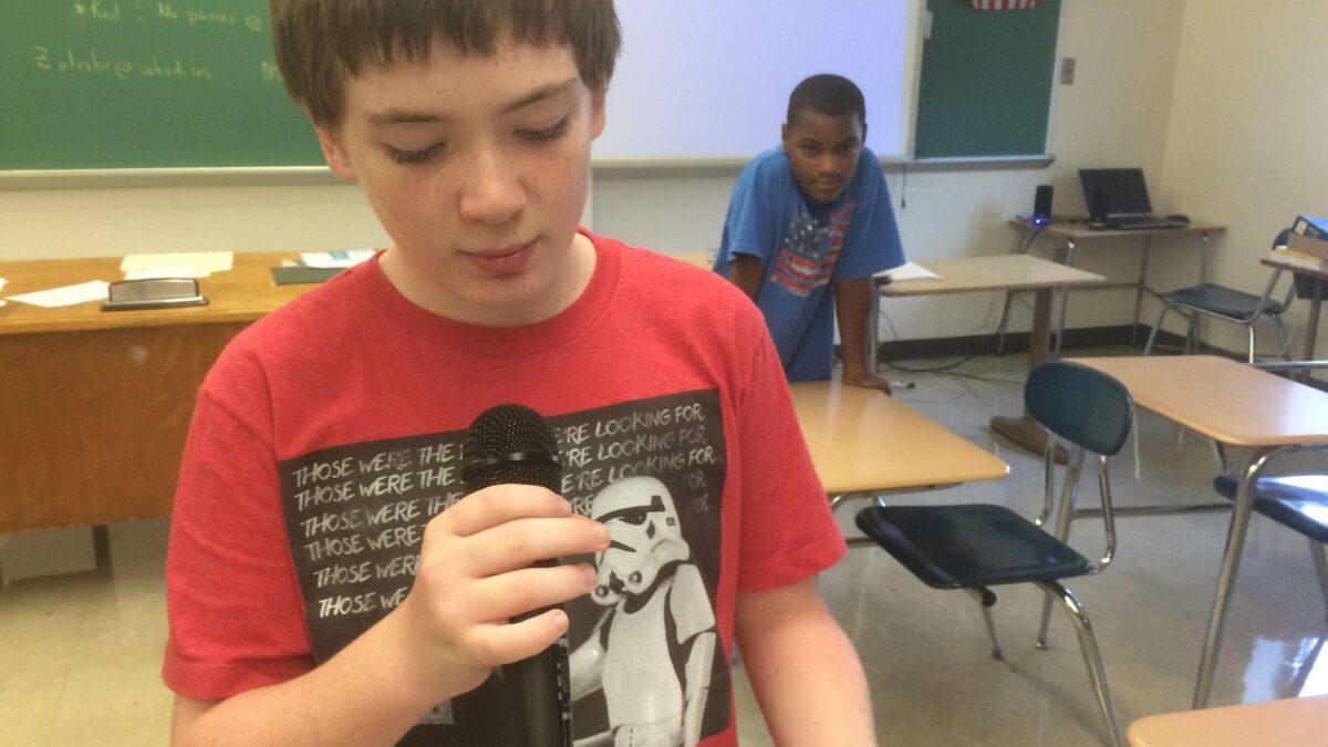 A youth holding and speaking into a microphone as another youth listens in a classroom