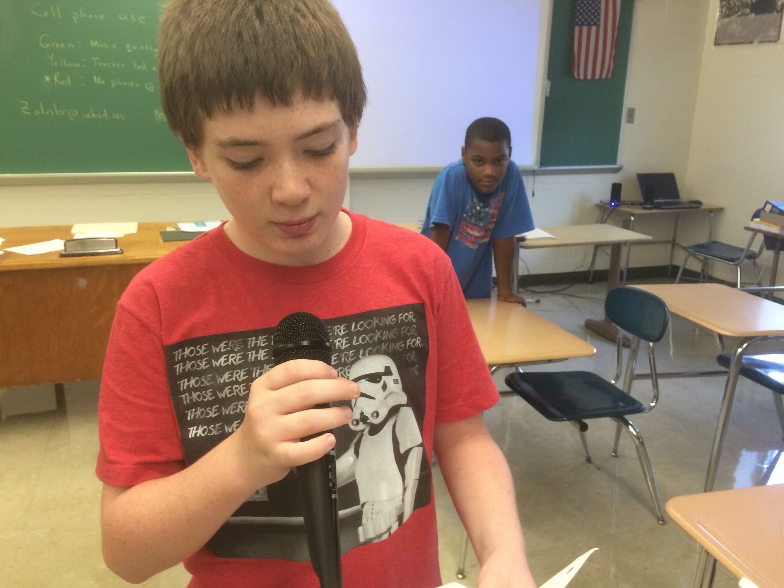 A youth holding and speaking into a microphone as another youth listens in a classroom