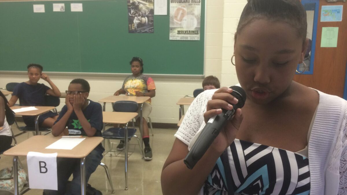 A youth holding a microphone as four youth listen sitting at classroom desks