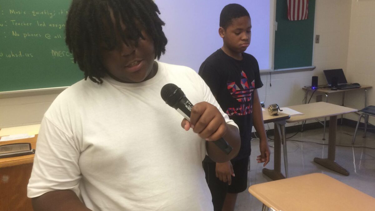 A youth holding a piece of paper speaking into a microphone as another youth listens in a classroom