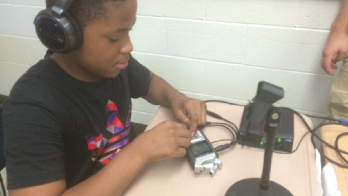 A youth wearing headphones interacting with a portable recorder on a classroom desk