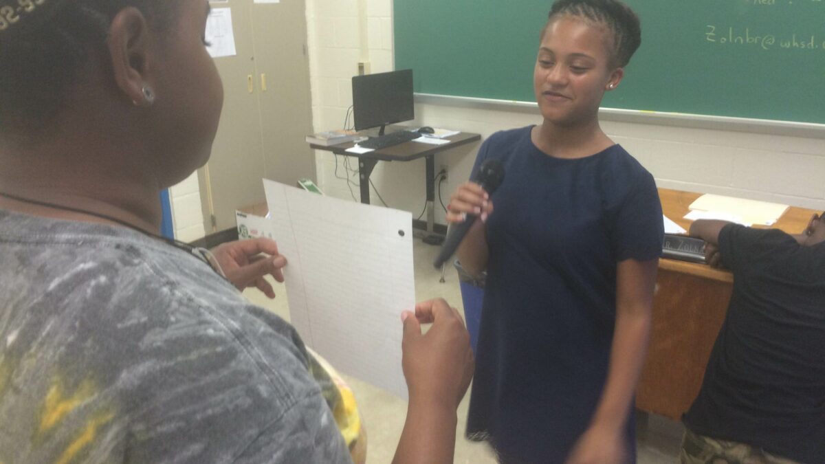 A youth holding up a piece of paper for another youth holding a microphone in a classroom