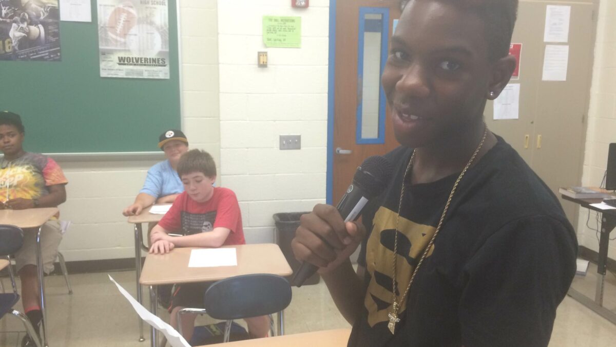 A youth holding a piece of paper and a microphone smiling at the camera as three youth listen sitting at classroom desks