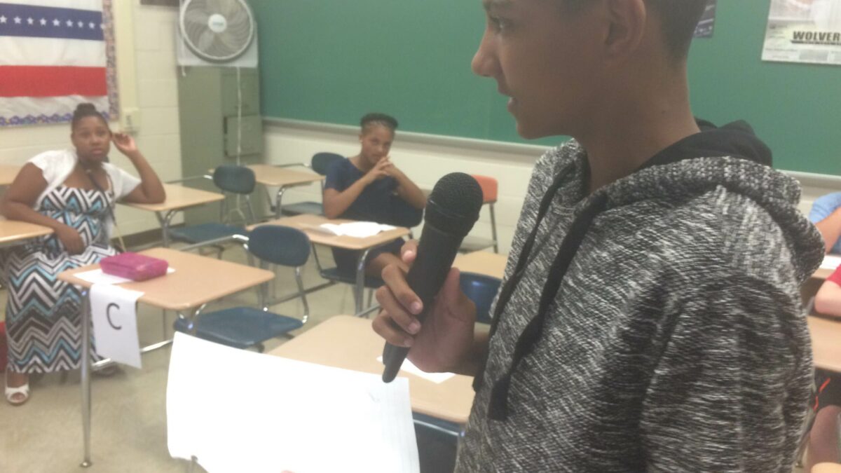 A youth holding a piece of paper speaking into a microphone as two youth listen sitting at classroom desks