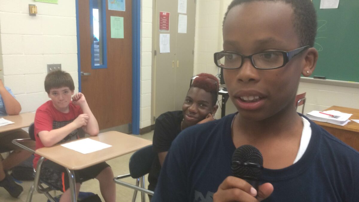 A youth holding a microphone as two youth listen sitting at classroom desks