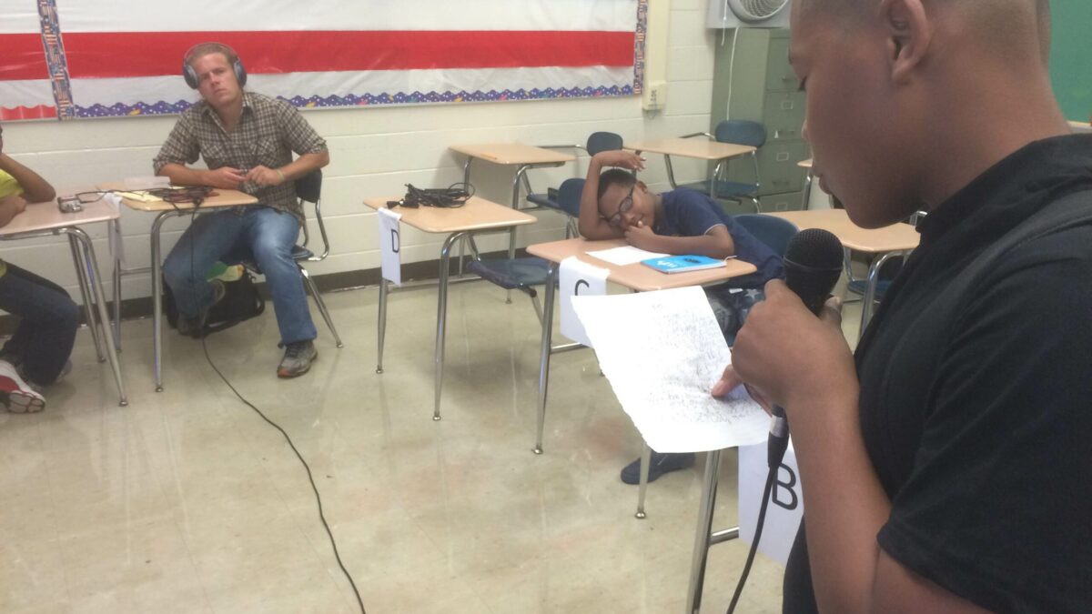 A youth holding a microphone reading a piece of paper in a classroom.