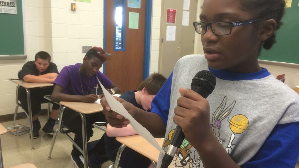 A youth reading a piece of paper and holding a microphone as three youth listen sitting at classroom desks