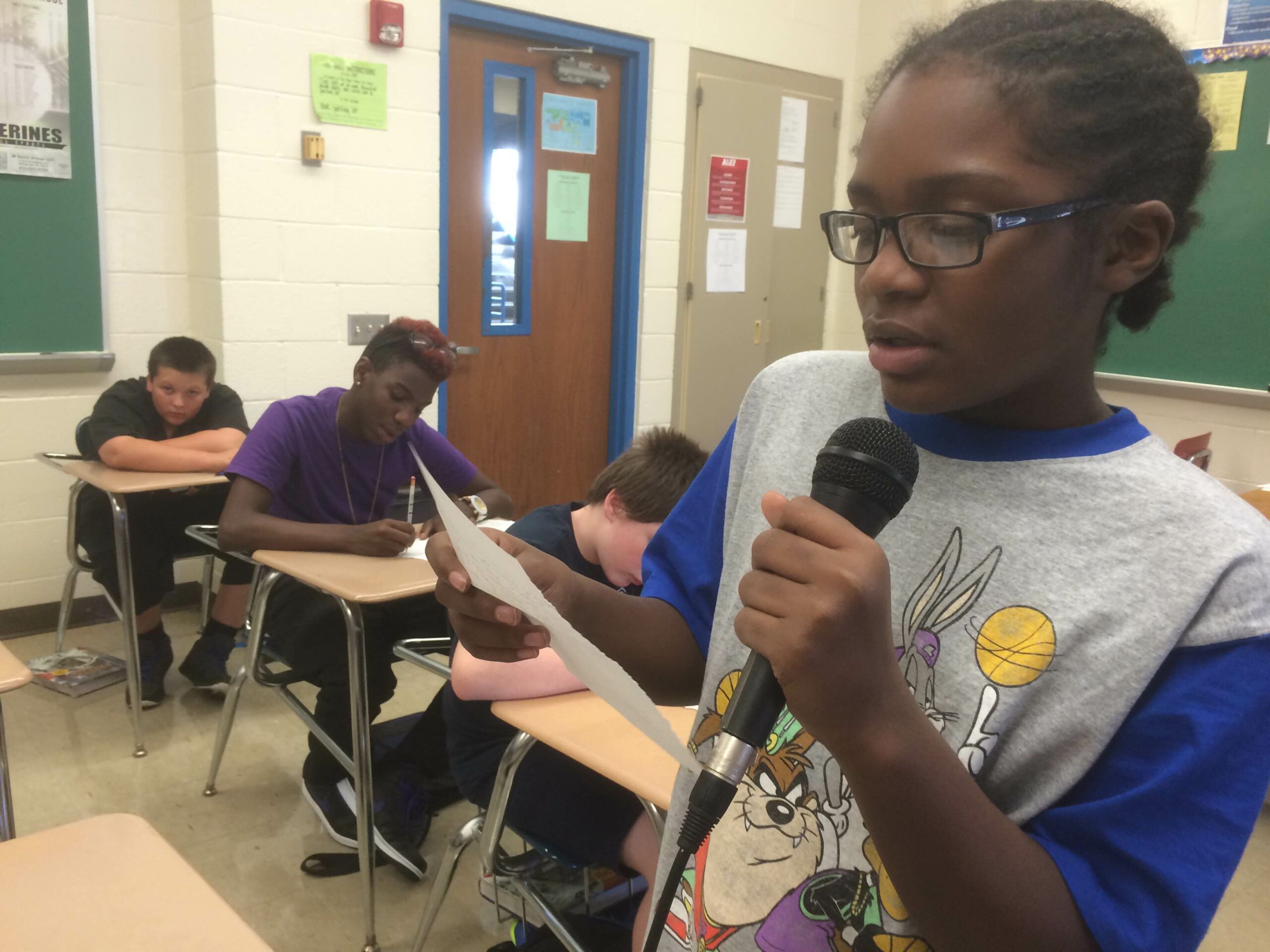 A youth reading a piece of paper and holding a microphone as three youth listen sitting at classroom desks