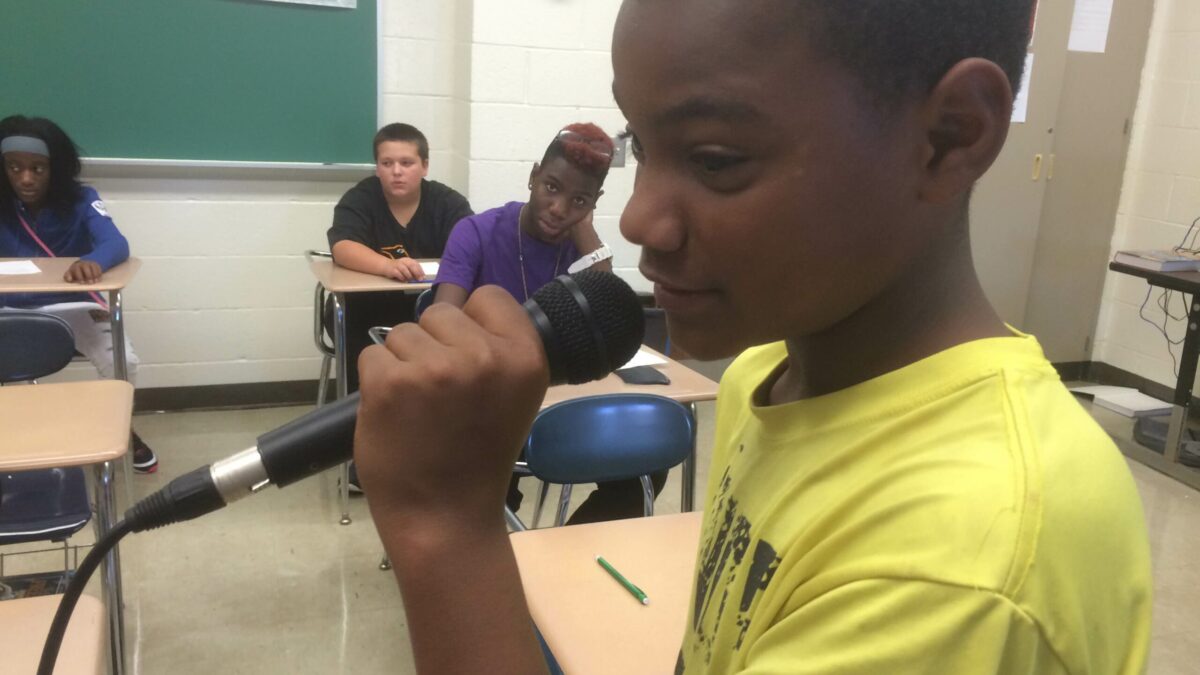 A youth holding a microphone as three youth listen sitting at classroom desks