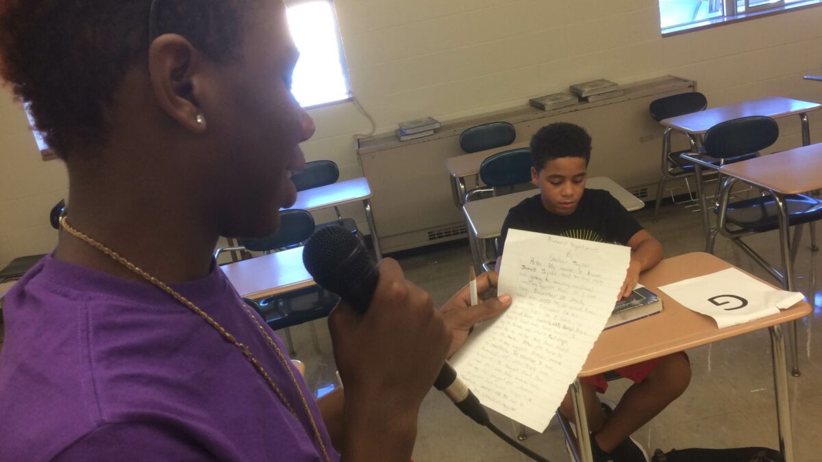 A youth holding a piece of paper and speaking into a microphone as another youth listens sitting at a classroom desk