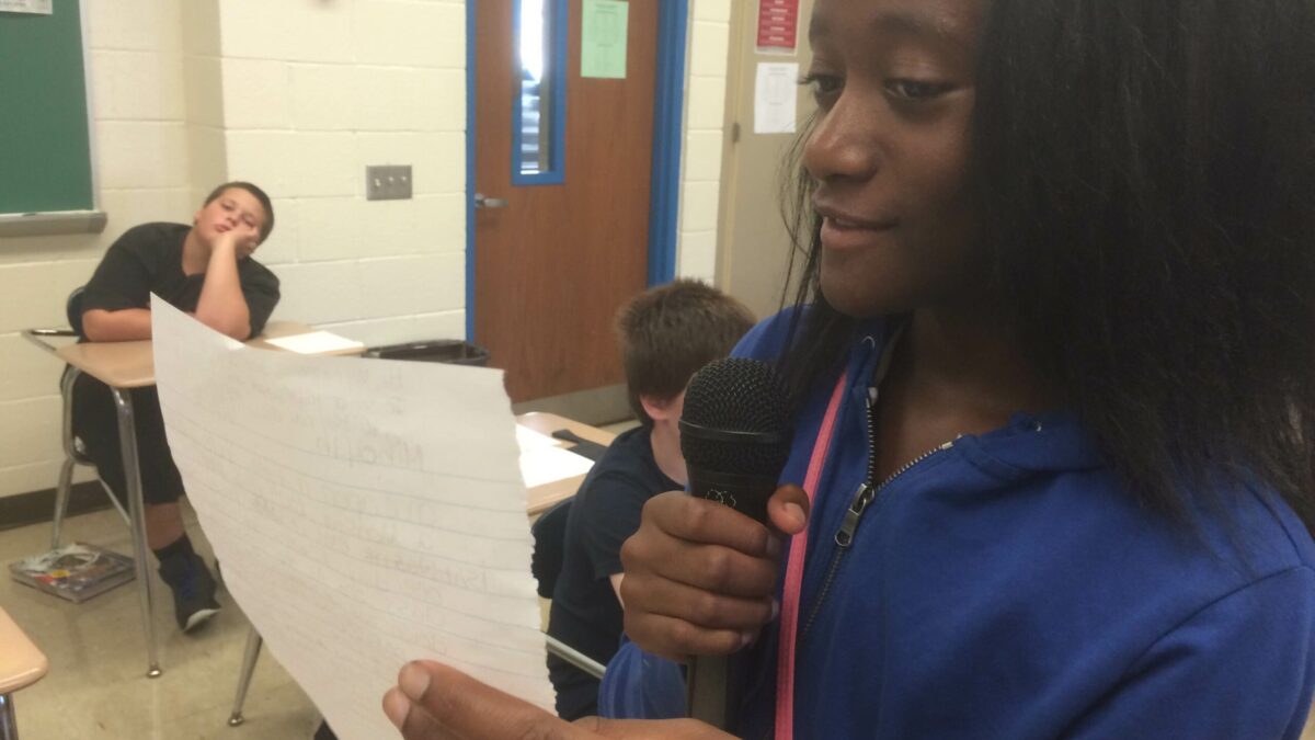 A youth holding a piece of paper and speaking into a microphone as two youths listen sitting at classroom desks
