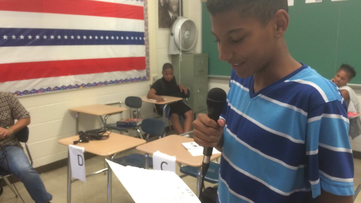 A youth holding a piece of paper and speaking into a microphone as three youth listen sitting at classroom desks