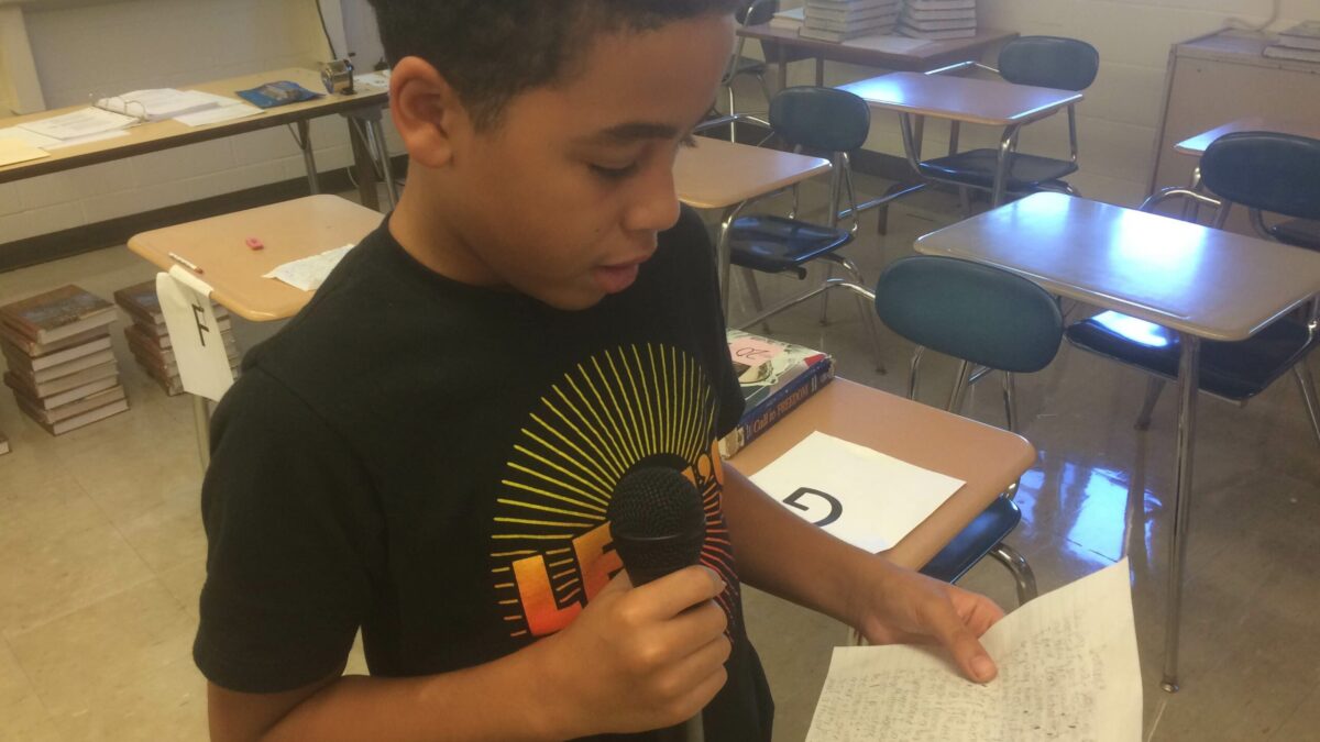 A youth holding a piece of paper and speaking into a microphone in a classroom