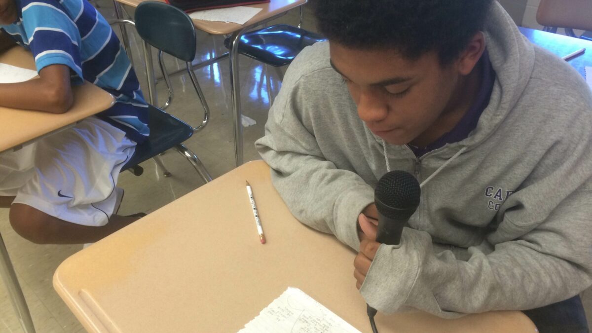A youth holding a microphone reading a piece of paper sitting on a classroom desk