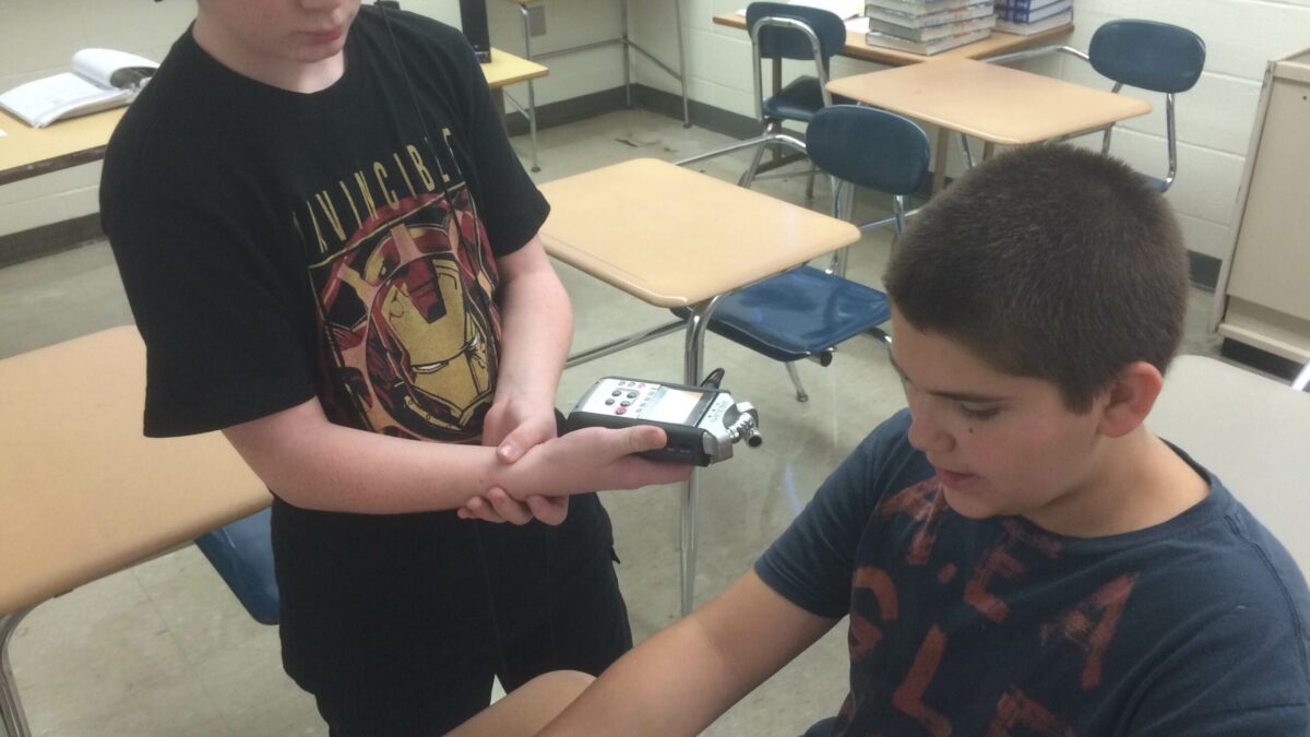 A youth wearing headphones holding up a portable recorder to another youth holding a pencil sitting at a classroom desk