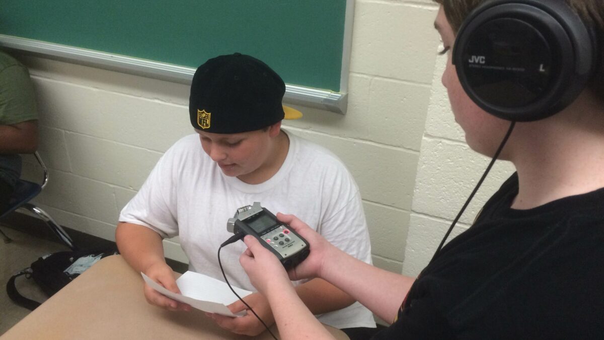 A youth wearing headphones holding up a portable recorder to another youth reading a piece of paper sitting at a classroom desk
