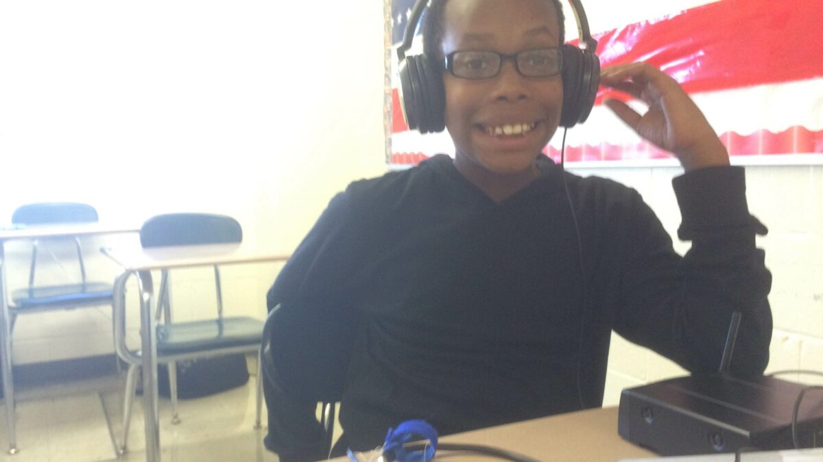 A youth wearing headphones connected to a portable recorder smiling sitting at a classroom desk