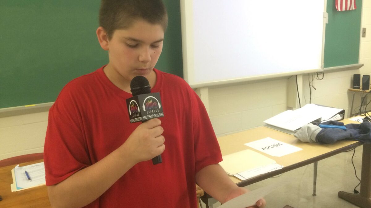 A youth holding a piece of paper and speaking into a microphone in a classroom