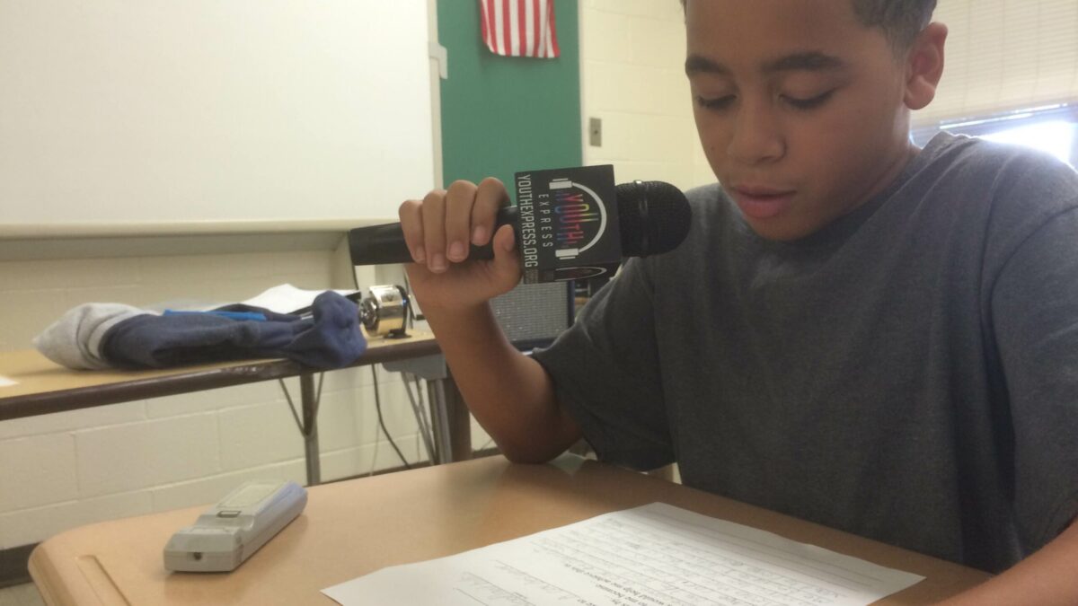 A youth holding a microphone looking down at a piece of paper on a classroom desk