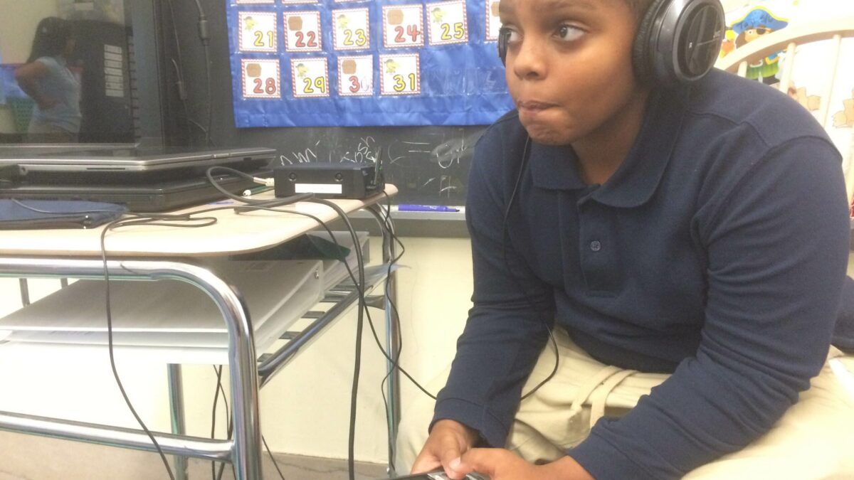 A youth wearing headphones holding a portable recorder sitting at a classroom desk
