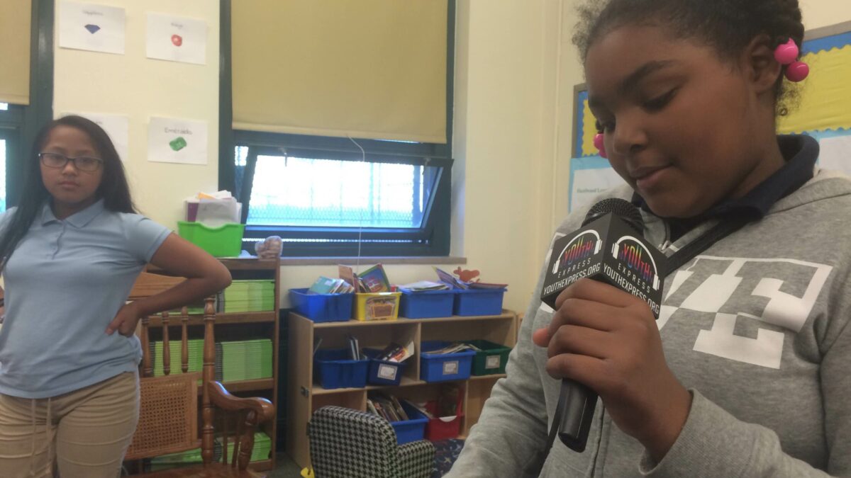 A youth looking at another youth holding a microphone and looking at a piece of paper in a classroom