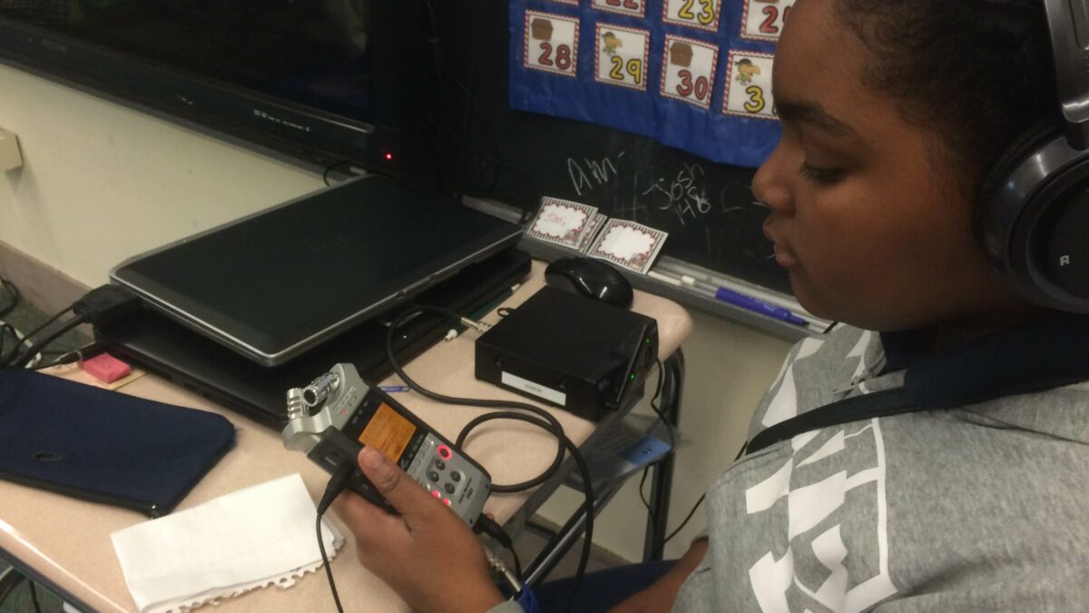 A youth wearing headphones holding a portable recorder sitting at a classroom desk