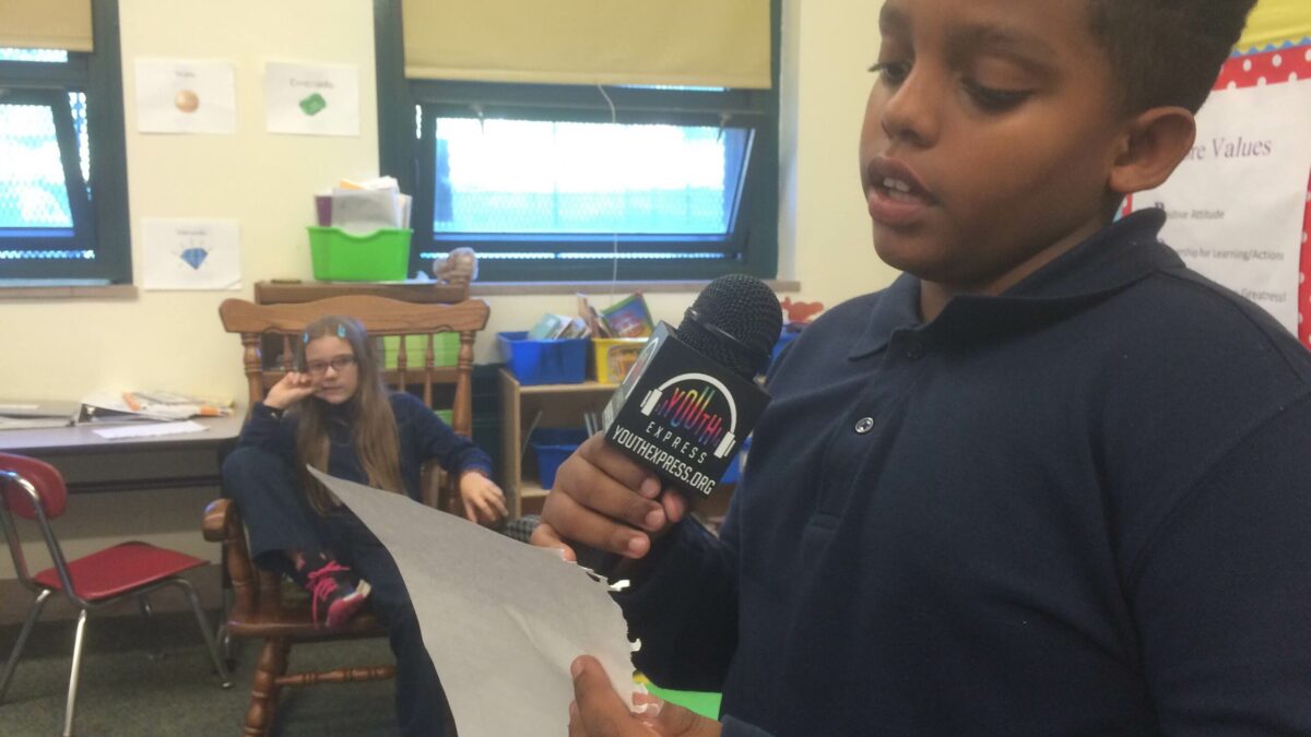 A youth looking at another youth holding a piece of paper and speaking into a microphone in a classroom