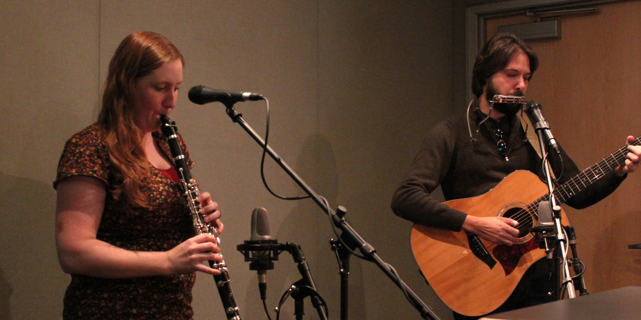 An adult playing clarinet and an adult playing acoustic guitar and playing a harmonica into a microphone in a radio studio