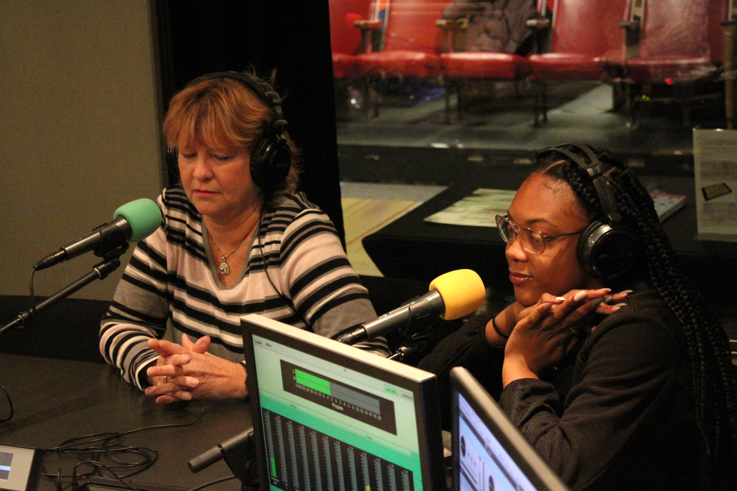 Two individuals wearing headphones sitting behind microphones in a radio studio