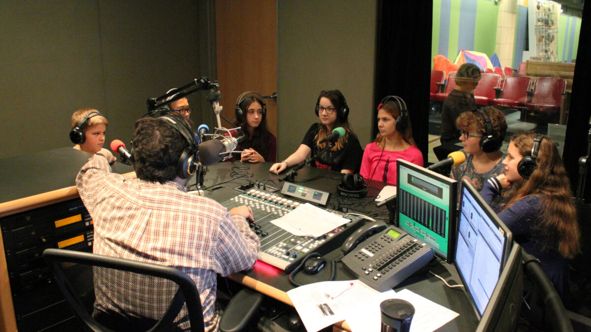 Seven youth wearing headphones sitting behind microphones being interviewed by an adult wearing headphones sitting behind a microphone and a mixing board in a radio studio