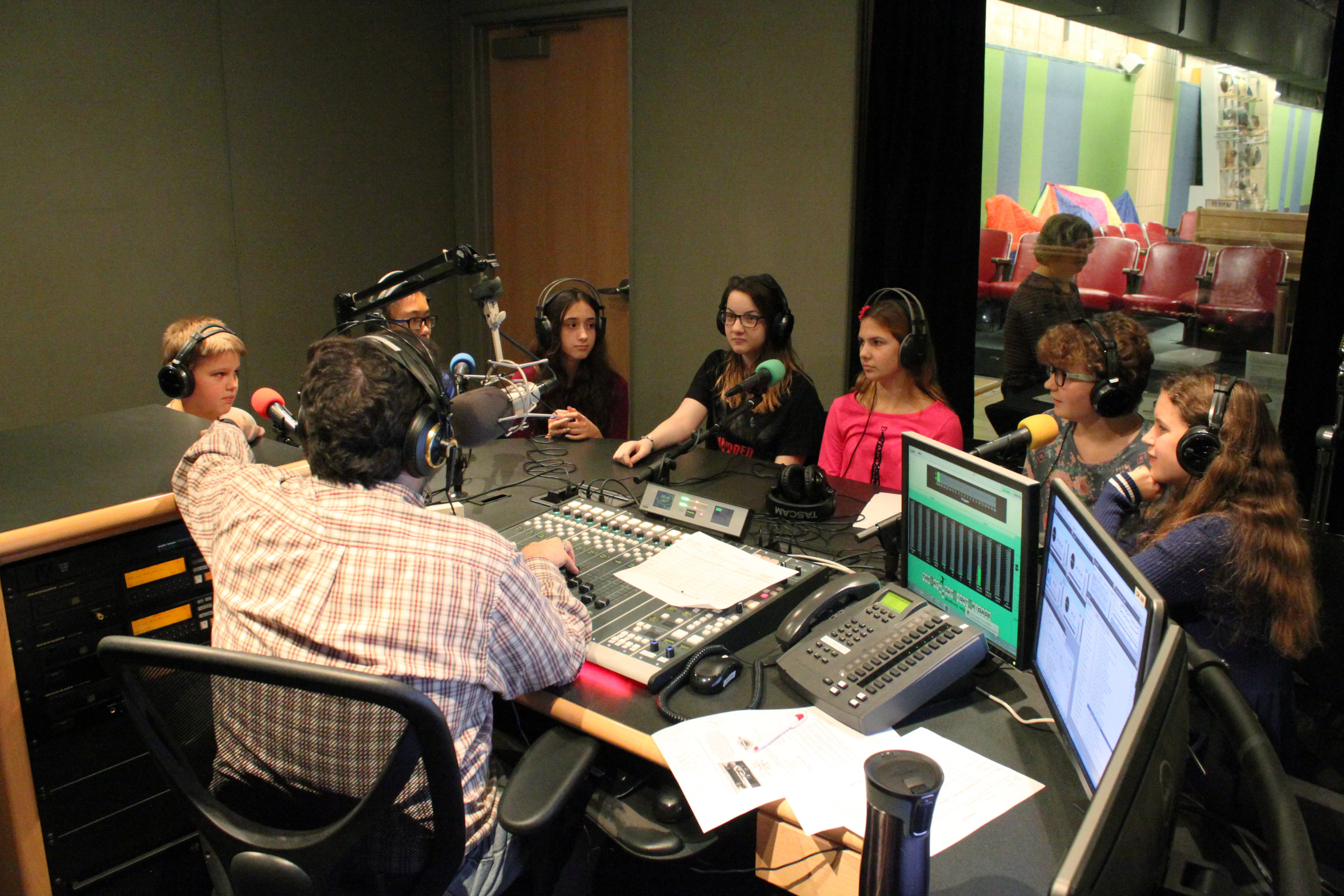 Seven youth wearing headphones sitting behind microphones being interviewed by an adult wearing headphones sitting behind a microphone and a mixing board in a radio studio
