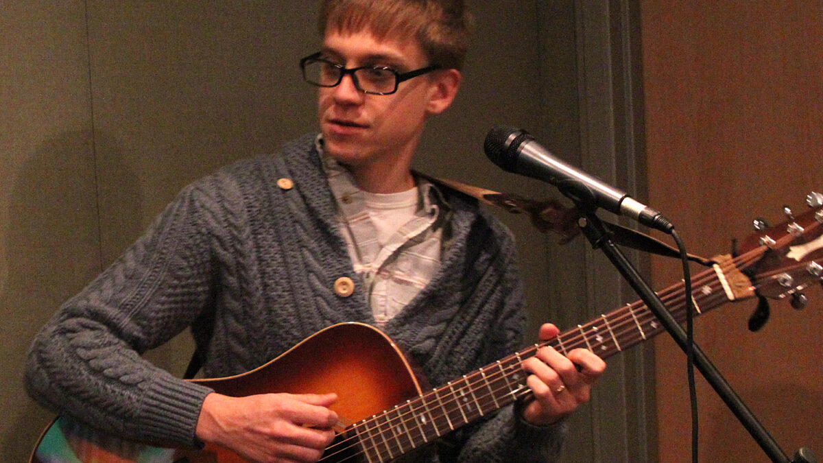 An adult playing acoustic guitar standing behind a microphone in a radio studio