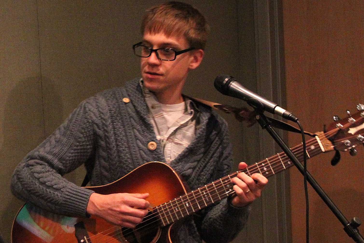 An adult playing acoustic guitar standing behind a microphone in a radio studio