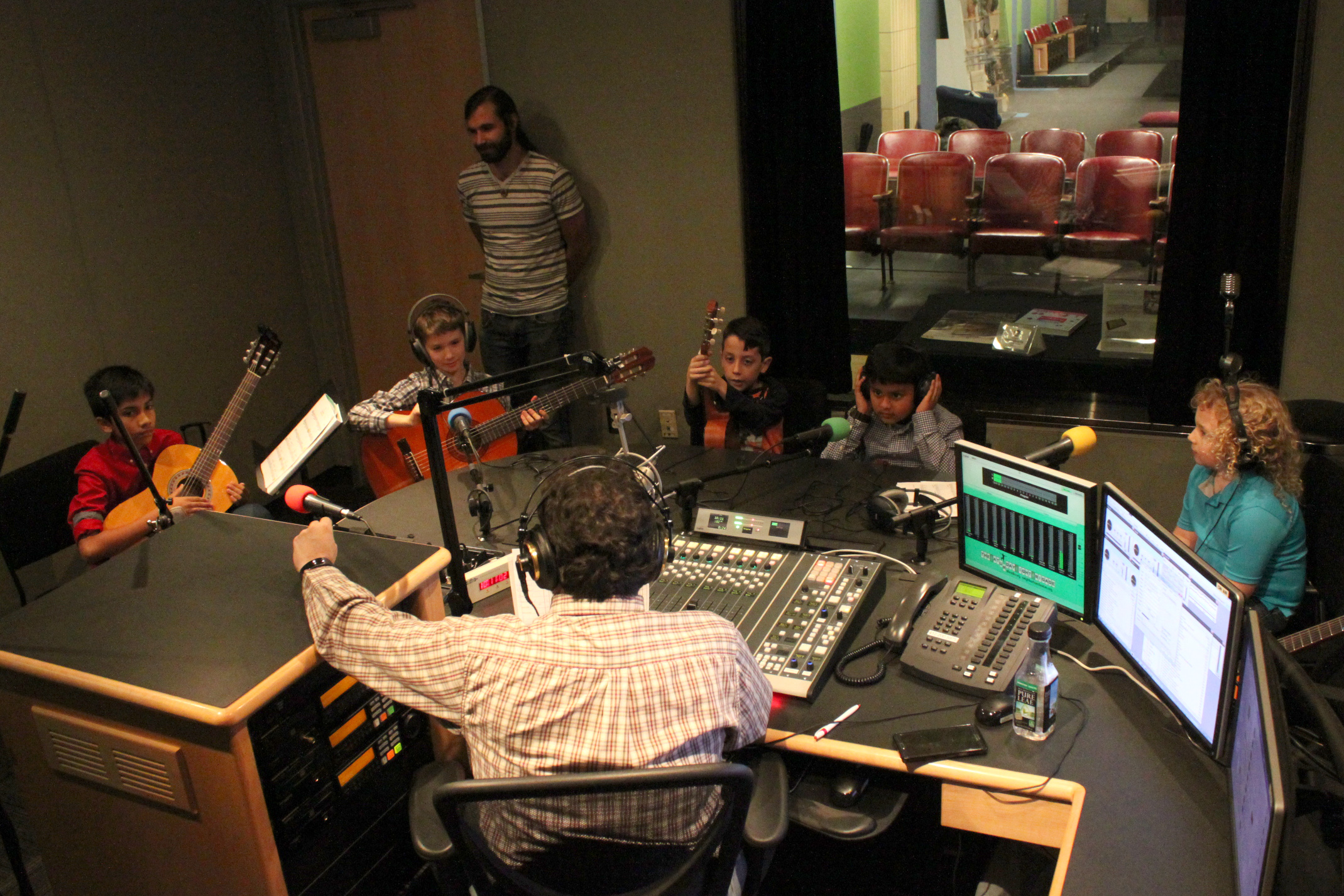 Five youth wearing headphones, three holding guitars, sitting behind microphones