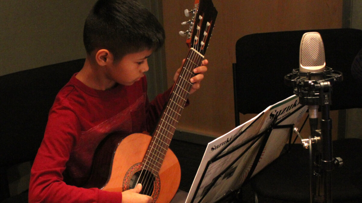 A youth playing acoustic guitar looking at sheet music on a music stand in a radio studio