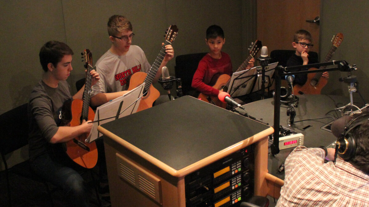 Four youth holding acoustic guitars being interviewed by an adult in a radio studio