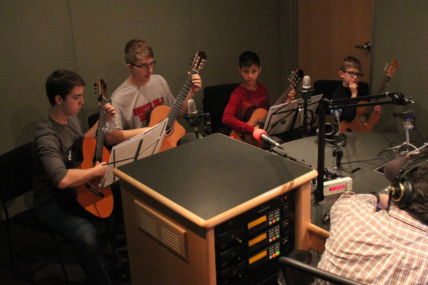 Four youth holding acoustic guitars being interviewed by an adult in a radio studio