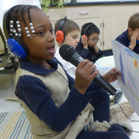 Three youth wearing headphones, one standing and holding and speaking into a microphone and holding a drawing on a piece of paper