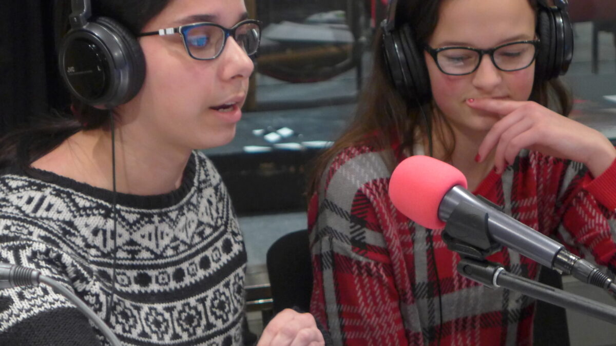 Two teens wearing headphones speaking into microphones in a radio studio