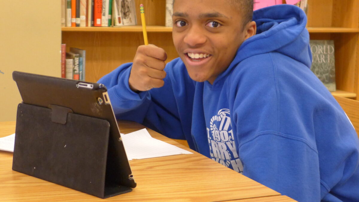 A teen sitting behind a piece of paper and an ipad smiling at the camera while holding a pencil in a library
