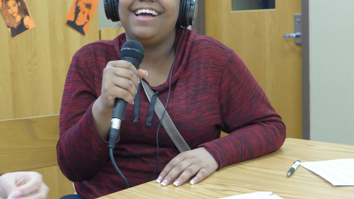 A teen wearing headphones and holding a microphone smiling while sitting behind a piece of paper on a brown table