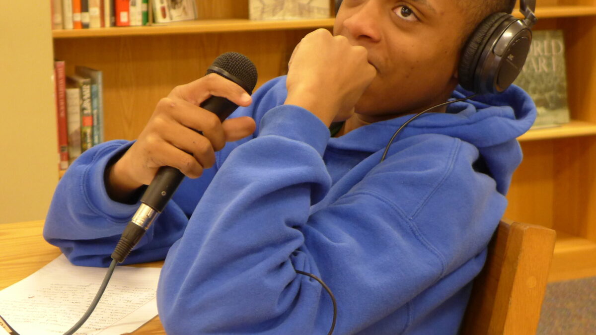 A teen wearing headphones and holding a microphone sitting behind a piece of paper on a brown table in a library