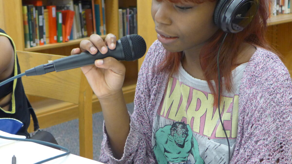 A teen wearing headphones sitting at a table holding a microphone in a library