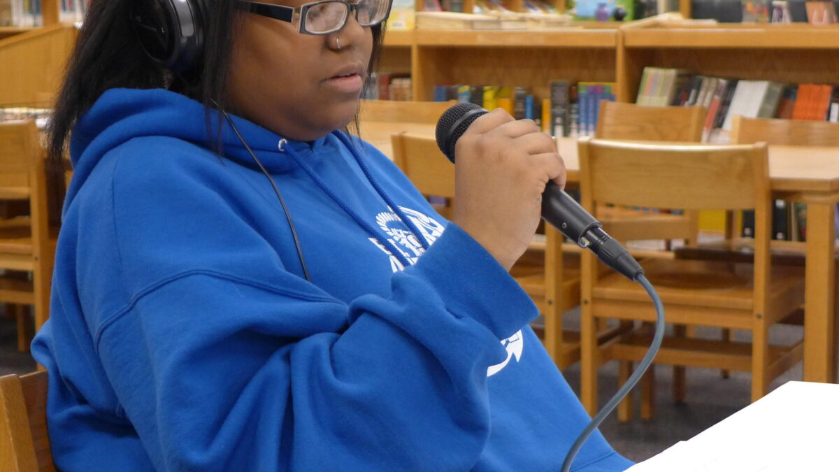A teen wearing headphones sitting at a table holding a microphone in a library